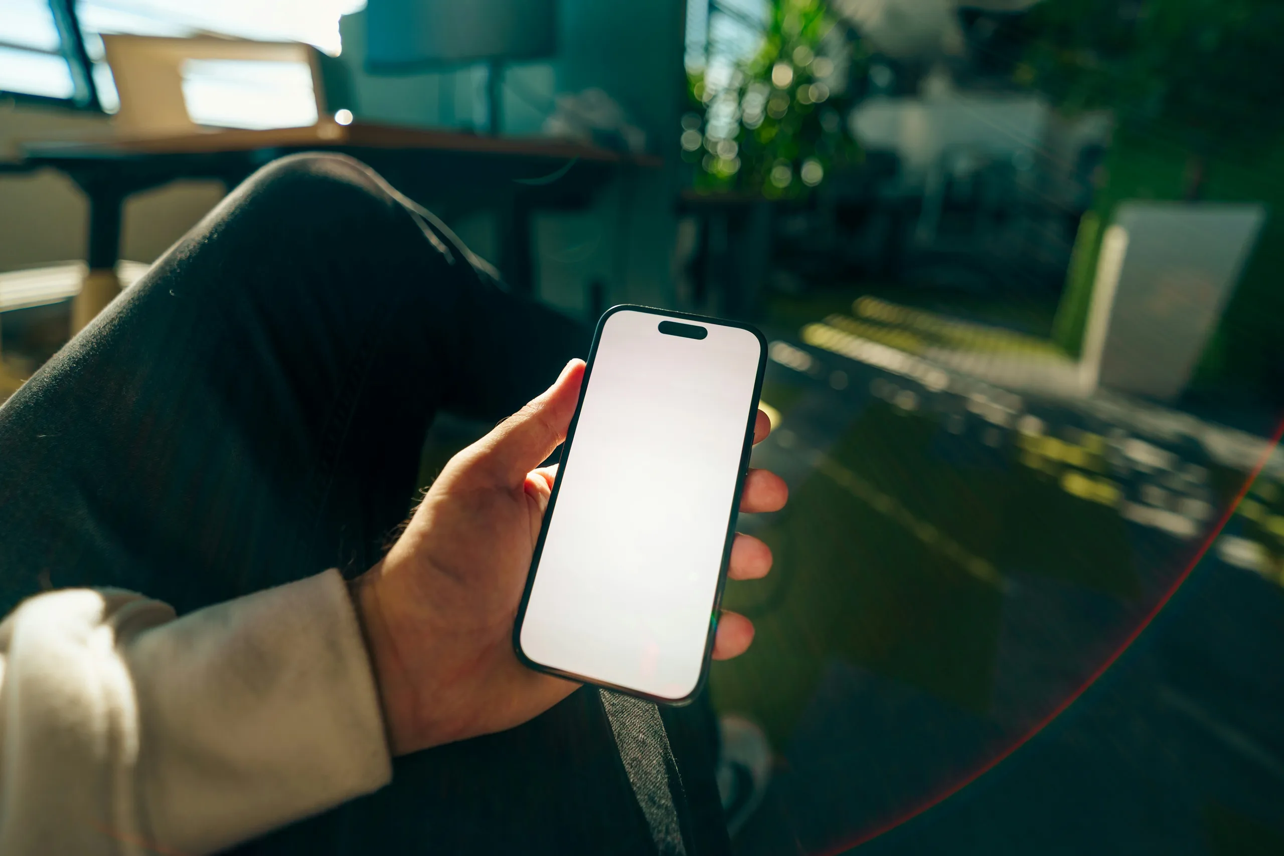 A hand holding a smartphone with a blank screen on a bright desk